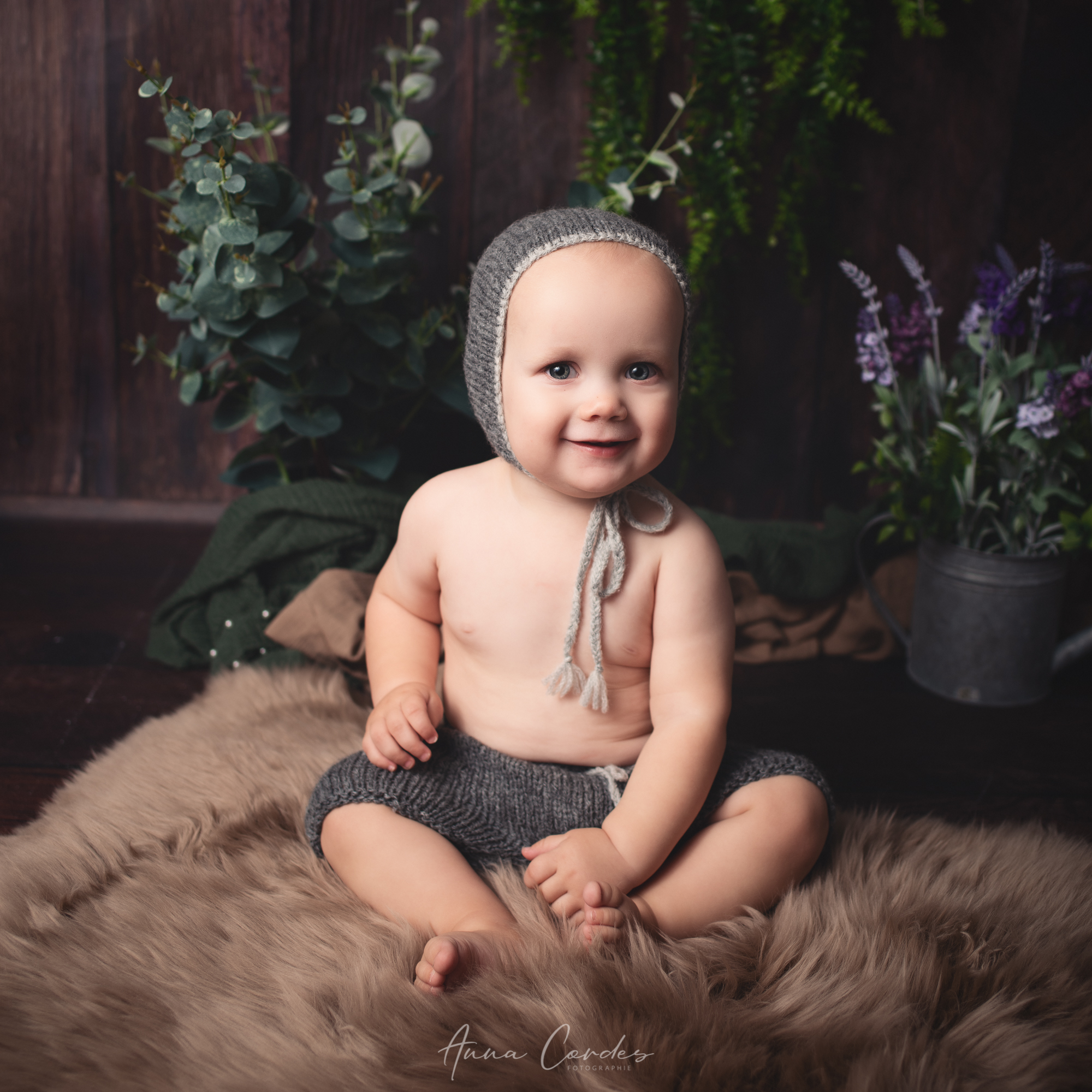 Baby mit Strickm&uuml;tze sitzt auf einem weichen Fell vor dunklem Hintergrund.