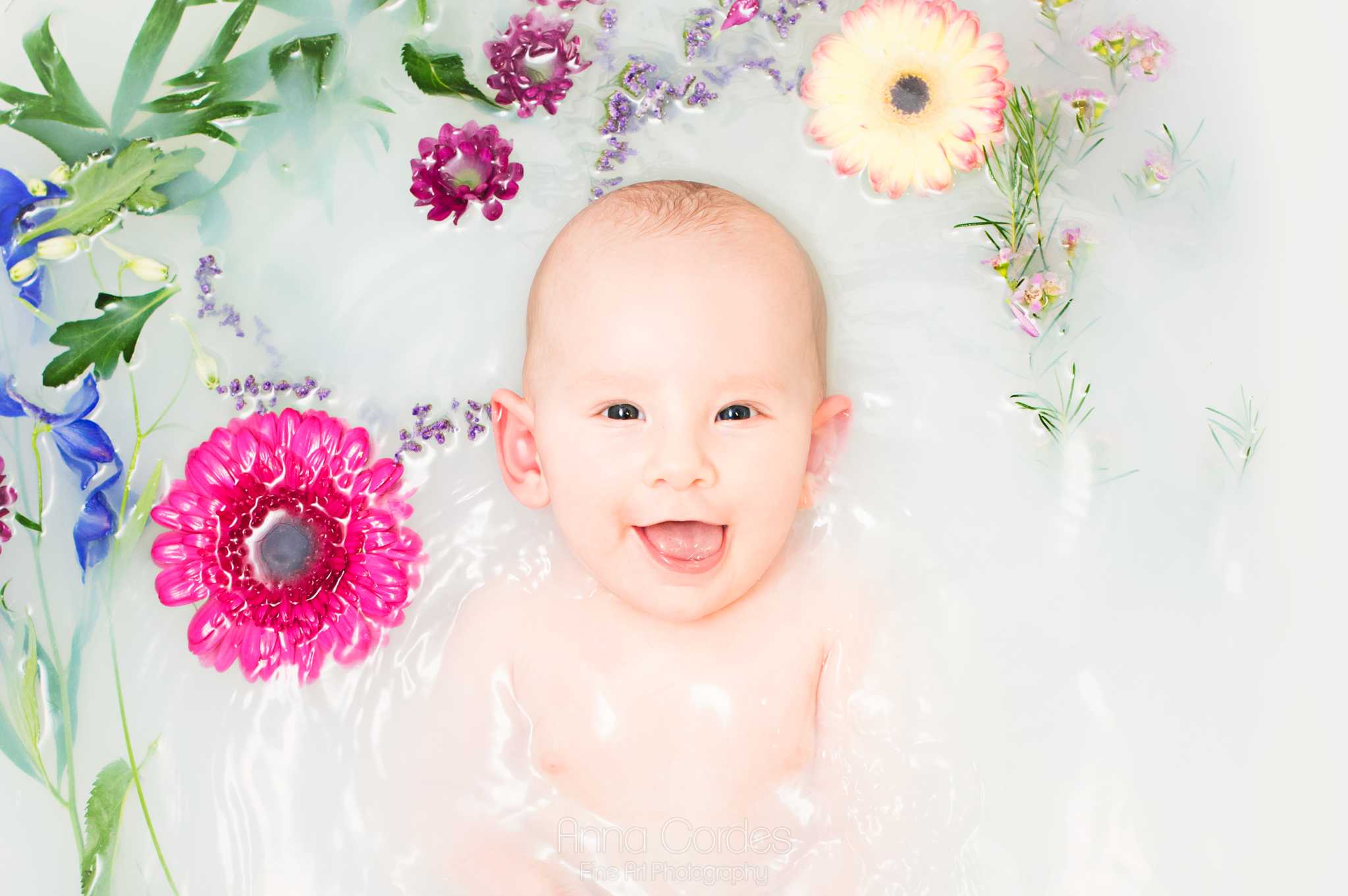 Baby sitzt lachend in einem Milchbad mit bunten Blumen.