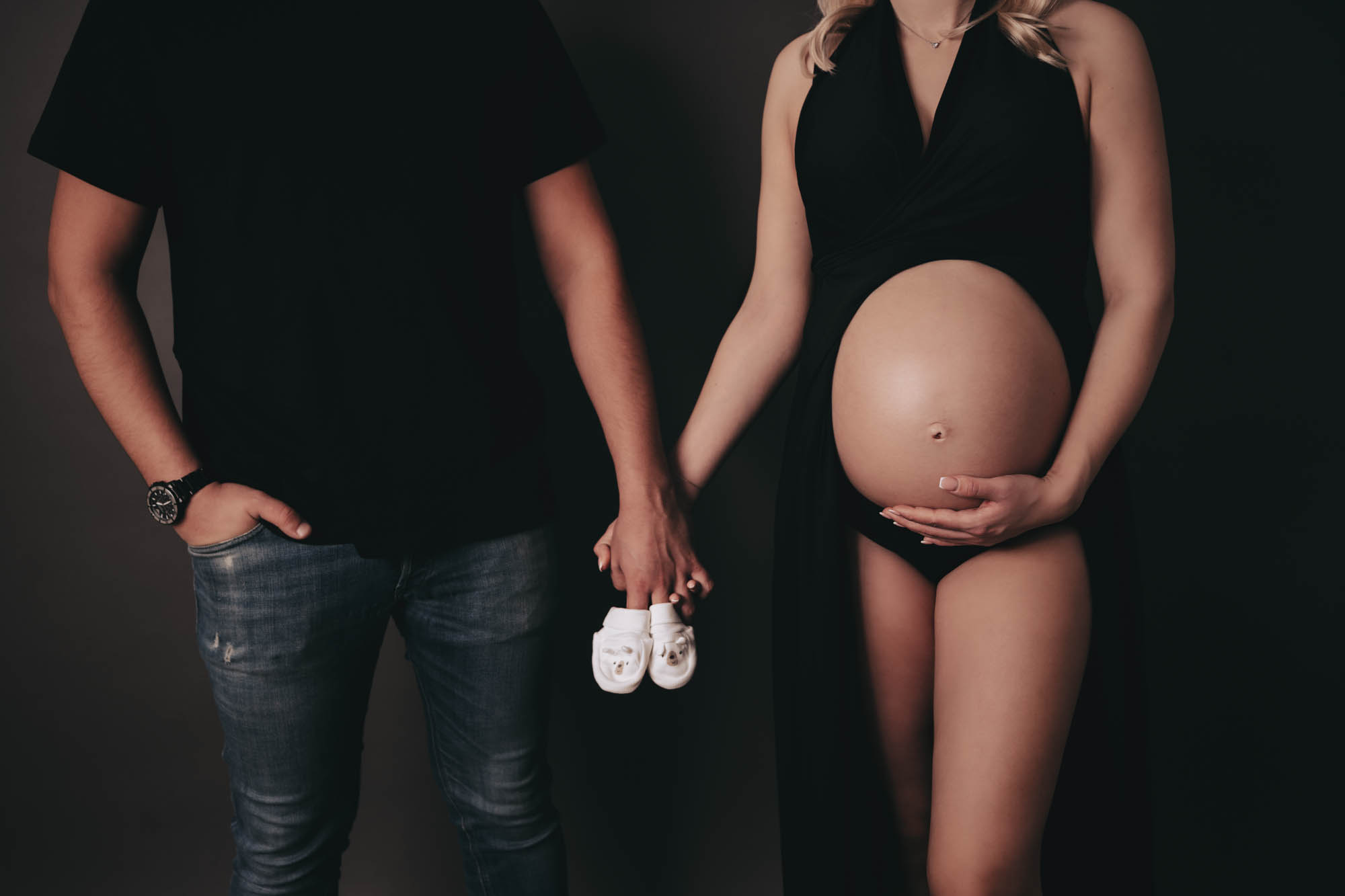 Elegantes Babybauchshooting Wien: Dunkles Paarfoto im schwarzen Kleid. Fokus auf nacktem Bauch, werdende Eltern Hand in Hand mit niedlichen Babyschuhen.