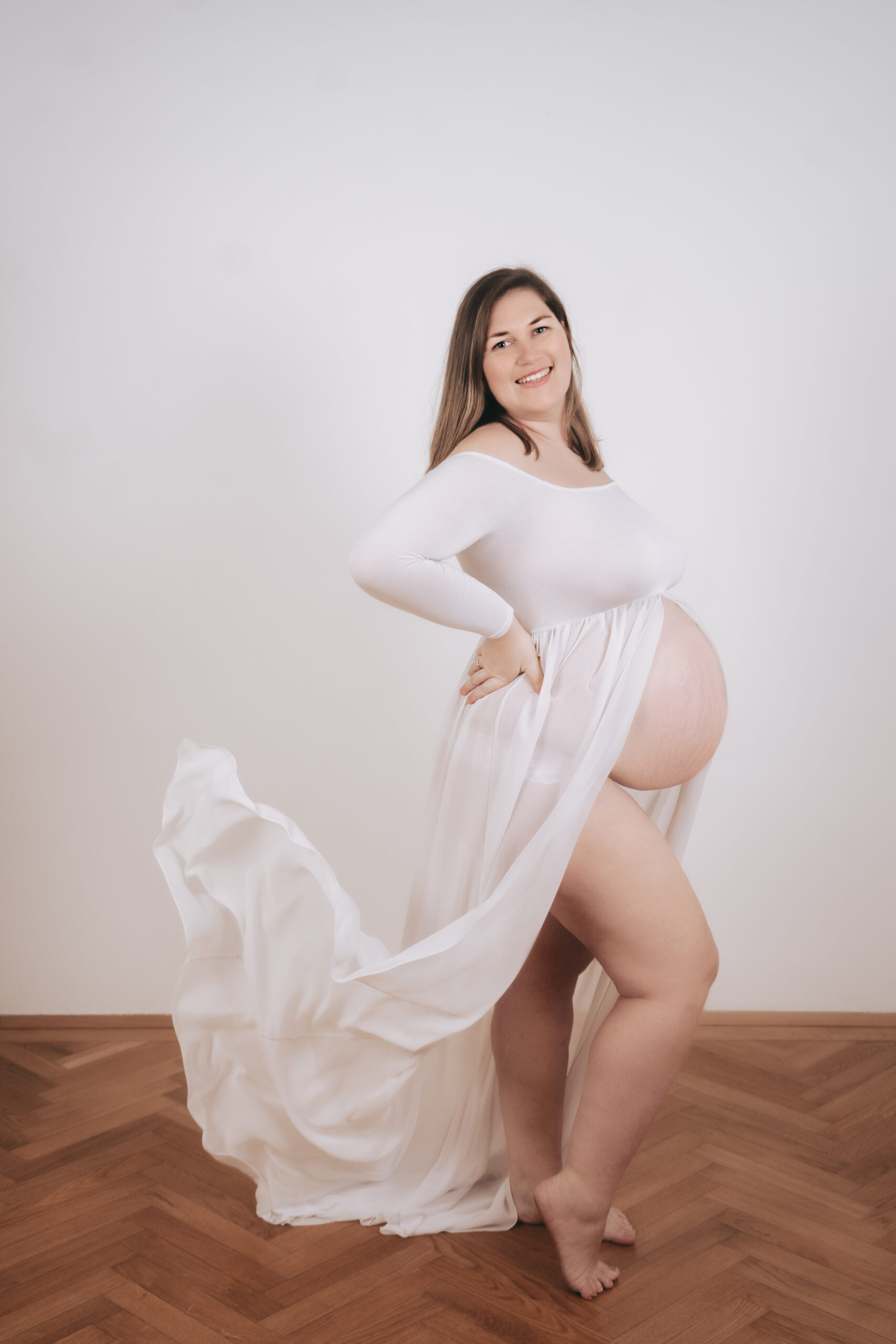Babybauchshooting Wien hell: Schwangere Frau in fließendem weißem Kleid mit nacktem Bauch im Studio.