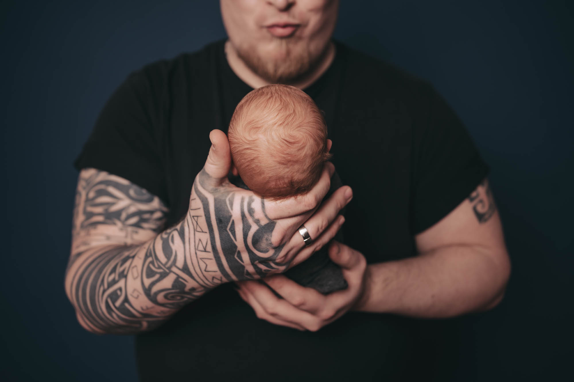 Neugeborenen Fotografie - T&auml;towierter Vater h&auml;lt sein Baby sch&uuml;tzend in den H&auml;nden.