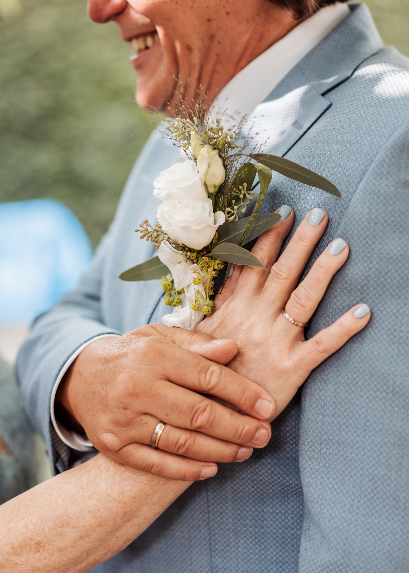 Nahaufnahme einer Hand mit Ehering, die auf dem Revers eines blauen Anzugs mit Ansteckblume liegt.